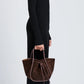 A person in a long black ribbed dress and boots stands in profile, holding the Proenza Schouler Small Ruched Soft Suede Tote with leather strap and bow detail, enhancing the sculptural silhouette of the ensemble.