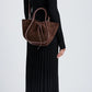 A woman in a long black ribbed dress and boots stands sideways, carrying the Proenza Schouler Small Ruched Soft Suede Tote with a sculptural silhouette over one shoulder.