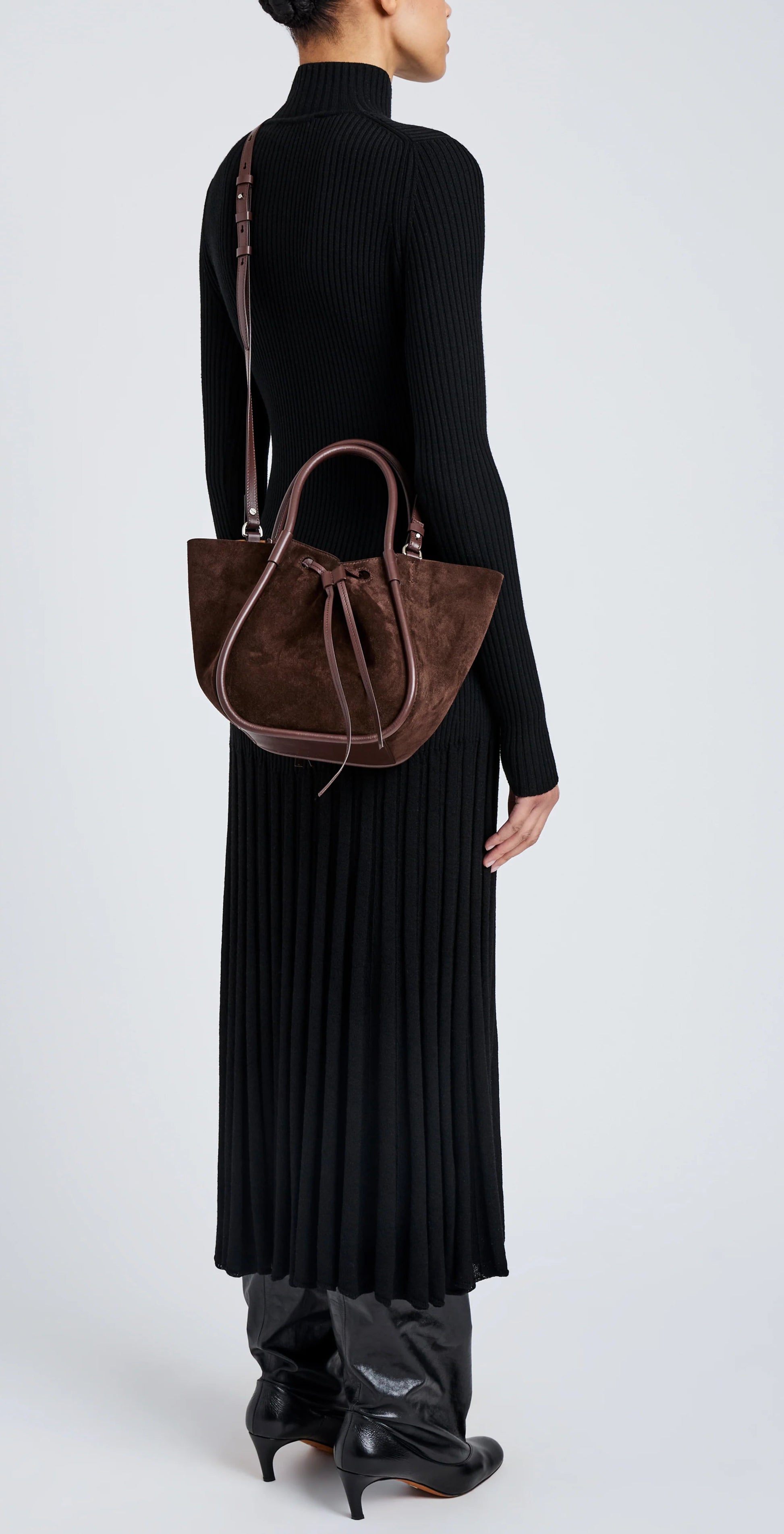 A woman in a long black ribbed dress and boots stands sideways, carrying the Proenza Schouler Small Ruched Soft Suede Tote with a sculptural silhouette over one shoulder.