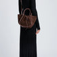 A person in a long black ribbed dress and boots stands against a plain light background, holding the Proenza Schouler Small Ruched Soft Suede Tote by its shoulder strap and handles.