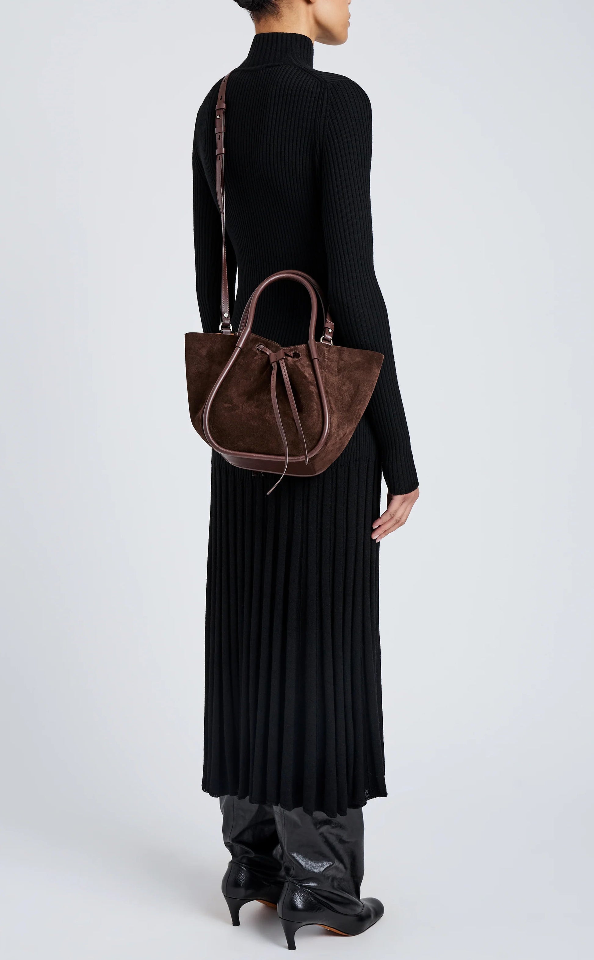 A person in a long black ribbed dress and boots stands against a plain light background, holding the Proenza Schouler Small Ruched Soft Suede Tote by its shoulder strap and handles.