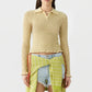 A woman wearing the Guest in Residence Smocked Long Sleeve Shrunken Polo in beige, paired with light denim shorts and a green patterned fabric tied around her waist, stands against a plain background.
