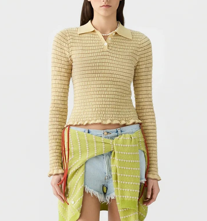 A woman wearing the Guest in Residence Smocked Long Sleeve Shrunken Polo in beige, paired with light denim shorts and a green patterned fabric tied around her waist, stands against a plain background.