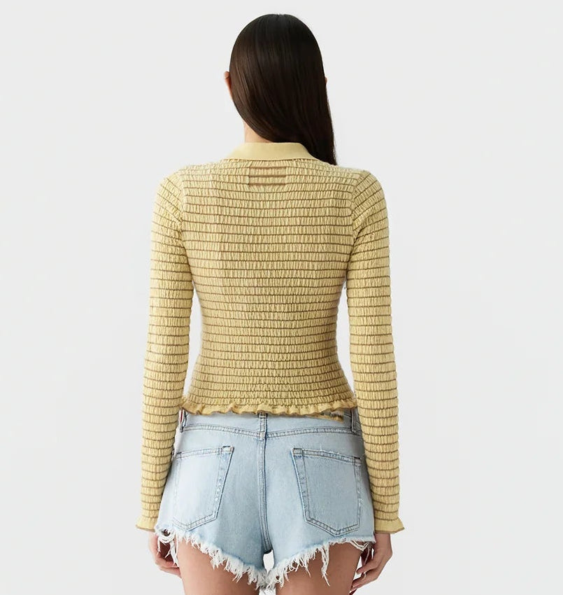 A person with long brown hair, wearing the Guest in Residence Smocked Long Sleeve Shrunken Polo in yellow and light blue frayed denim shorts, stands with their back to the camera against a plain light background.