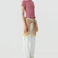 A person in a slim fit Smocked Shrunken Polo by Guest in Residence, pink short-sleeve knit top, white pants, light sneakers, and a striped sweater tied at the waist stands against a plain light background.