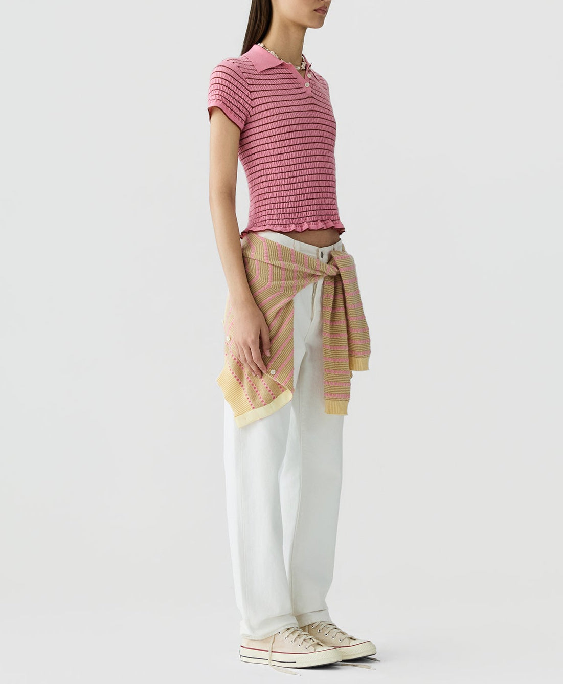A person in a slim fit Smocked Shrunken Polo by Guest in Residence, pink short-sleeve knit top, white pants, light sneakers, and a striped sweater tied at the waist stands against a plain light background.