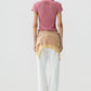 A person stands with their back to the camera against a plain background, wearing the Guest in Residence Smocked Shrunken Polo in pink, white pants, and a yellow sweater tied around their waist.