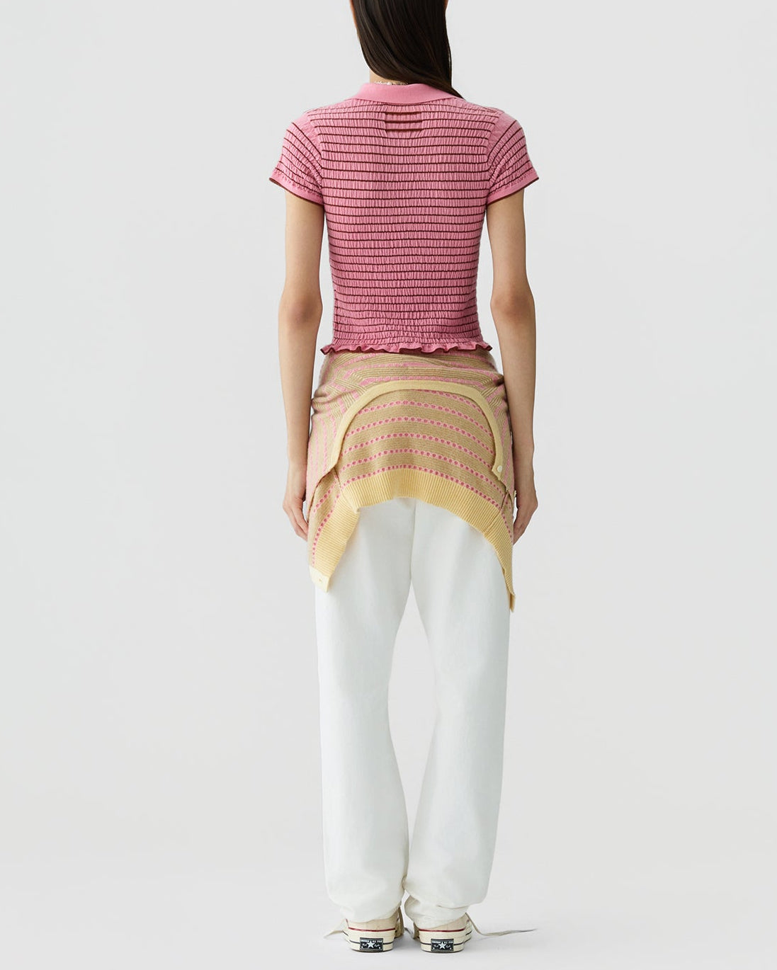 A person stands with their back to the camera against a plain background, wearing the Guest in Residence Smocked Shrunken Polo in pink, white pants, and a yellow sweater tied around their waist.