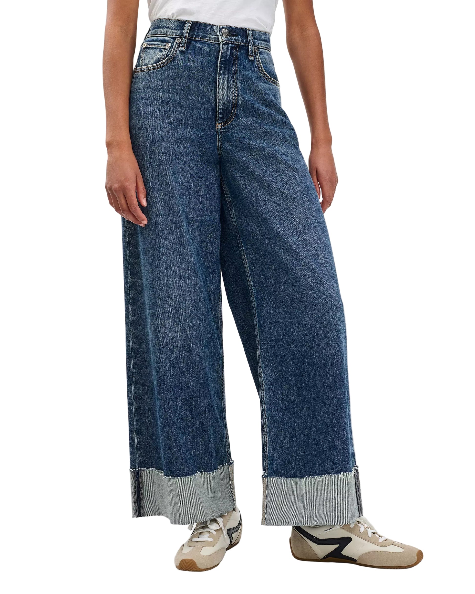 A person wearing Rag & Bone Sofie High Rise Full Length Cuff vintage stretch denim wide-leg blue jeans with frayed, cuffed hems, paired with a white shirt and beige and black sneakers.