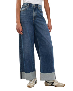 A person wearing Rag & Bone Sofie High Rise Full Length Cuff vintage stretch denim wide-leg blue jeans with frayed, cuffed hems, paired with a white shirt and beige and black sneakers.