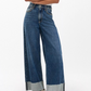 A person stands with arms crossed, wearing Rag & Bone's Sofie High Rise Full Length Cuff jeans paired with a white T-shirt and beige and black sneakers, set against a plain background.