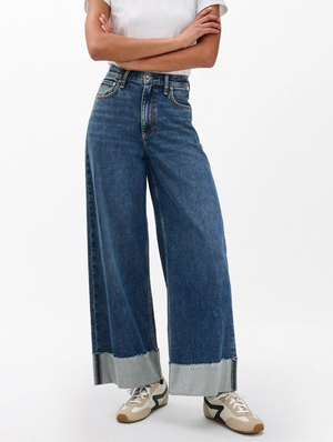 A person stands with arms crossed, wearing Rag & Bone's Sofie High Rise Full Length Cuff jeans paired with a white T-shirt and beige and black sneakers, set against a plain background.