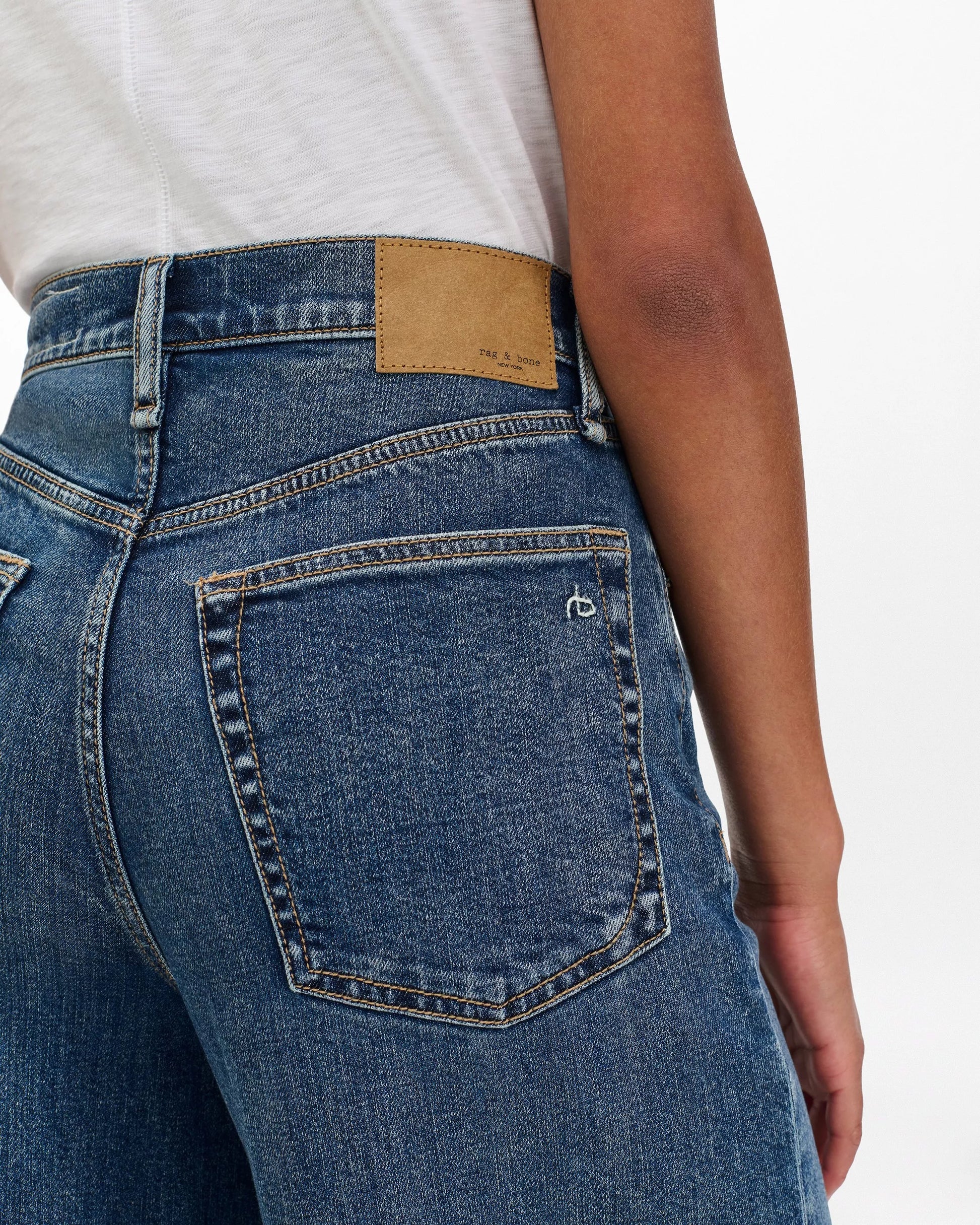 A person shown from the back wears Rag & Bone's Sofie High Rise Full Length Cuff jeans in blue denim and a white shirt. The wide leg jeans feature a large back pocket with subtle stitching and a tan leather patch on the waistband.