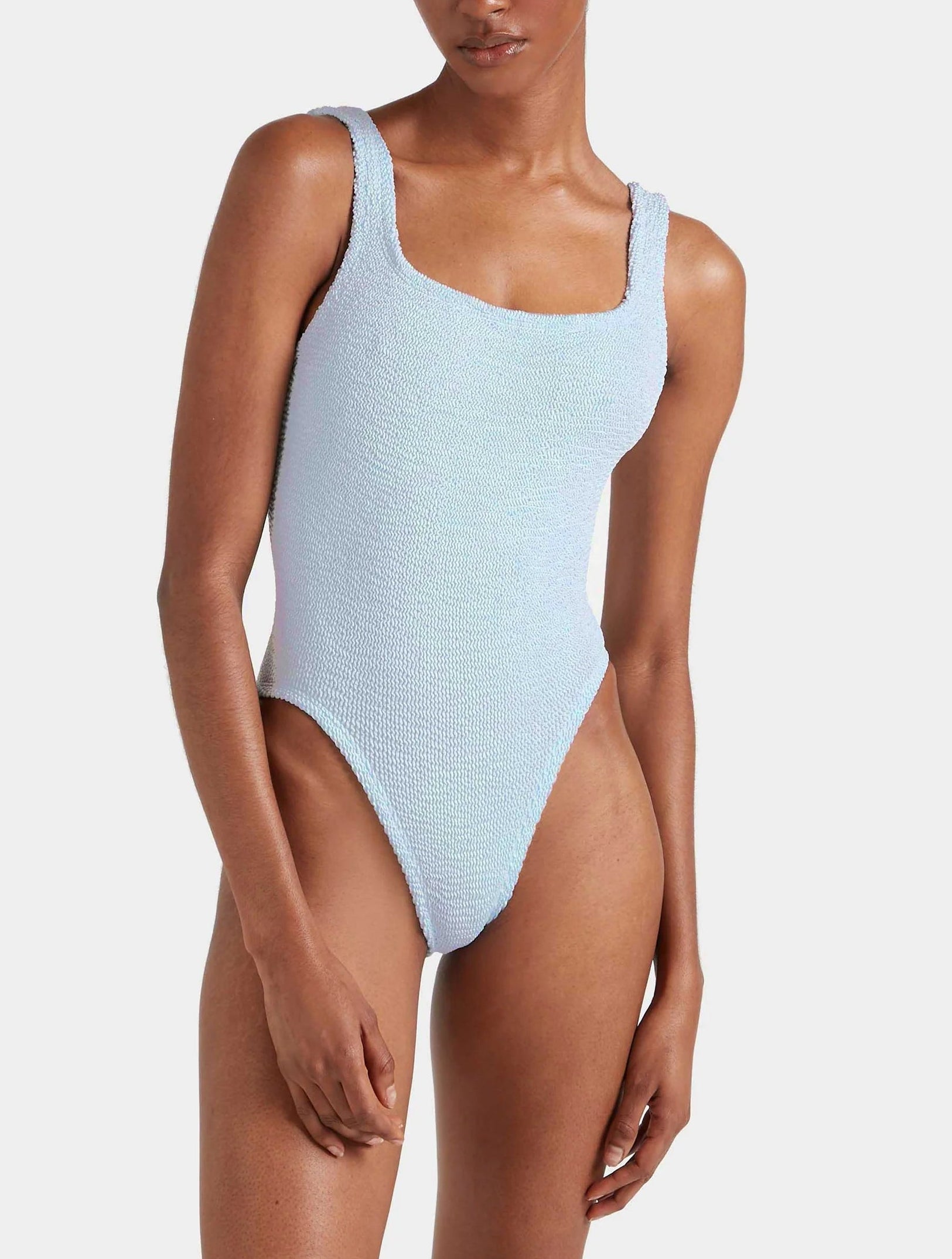 Model wears the Hunza G Square Neck Swim, a light blue crinkle fabric one-piece with high-cut legs and wide straps, posed against a plain white background.