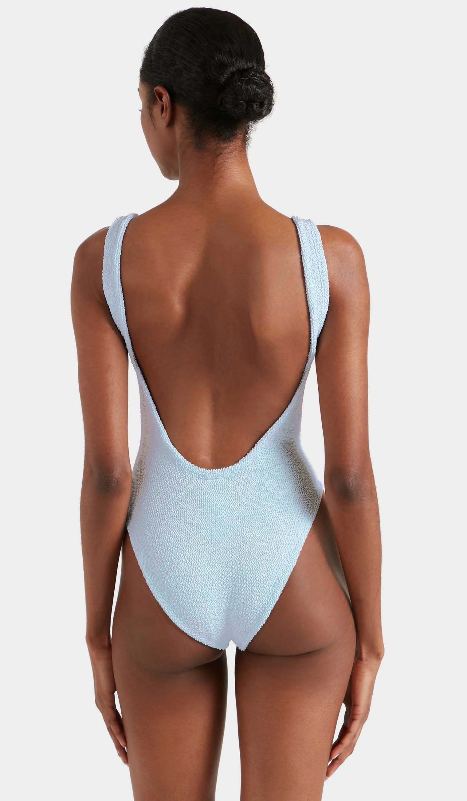 A person faces away from the camera, wearing the Hunza G Square Neck Swim in light blue. Made from signature crinkle fabric, this one-size swimsuit features a square neckline, low scoop back, and high-cut legs for a flattering fit.