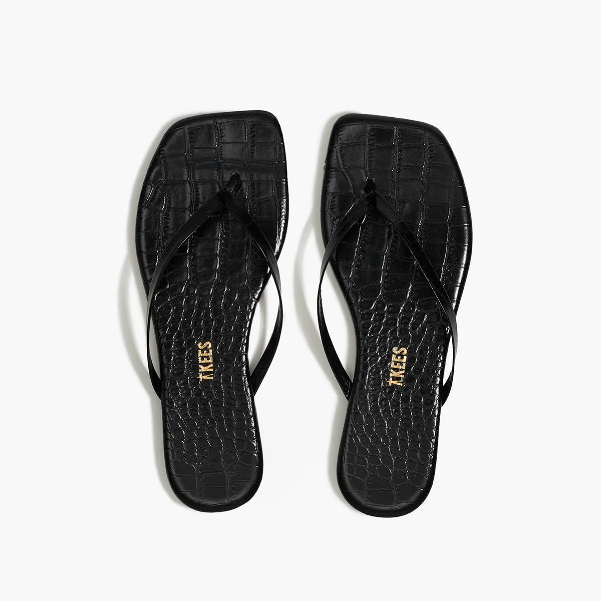 The Lily Square Toe Croc Sandal by Tkees features a square toe, thin straps, and black crocodile-embossed leather. The insole is adorned with the Tkees brand name in gold lettering for an elegant touch.
