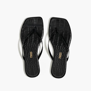 The Lily Square Toe Croc Sandal by Tkees features a square toe, thin straps, and black crocodile-embossed leather. The insole is adorned with the Tkees brand name in gold lettering for an elegant touch.
