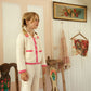 A young child with blonde hair in braids stands on a chair in a vintage-style room with floral décor and white paneled walls, wearing the Girls’ Josia Cardigan by Louise Misha, a cozy cotton knit sweater adorned with delicate hand-crocheted flowers.