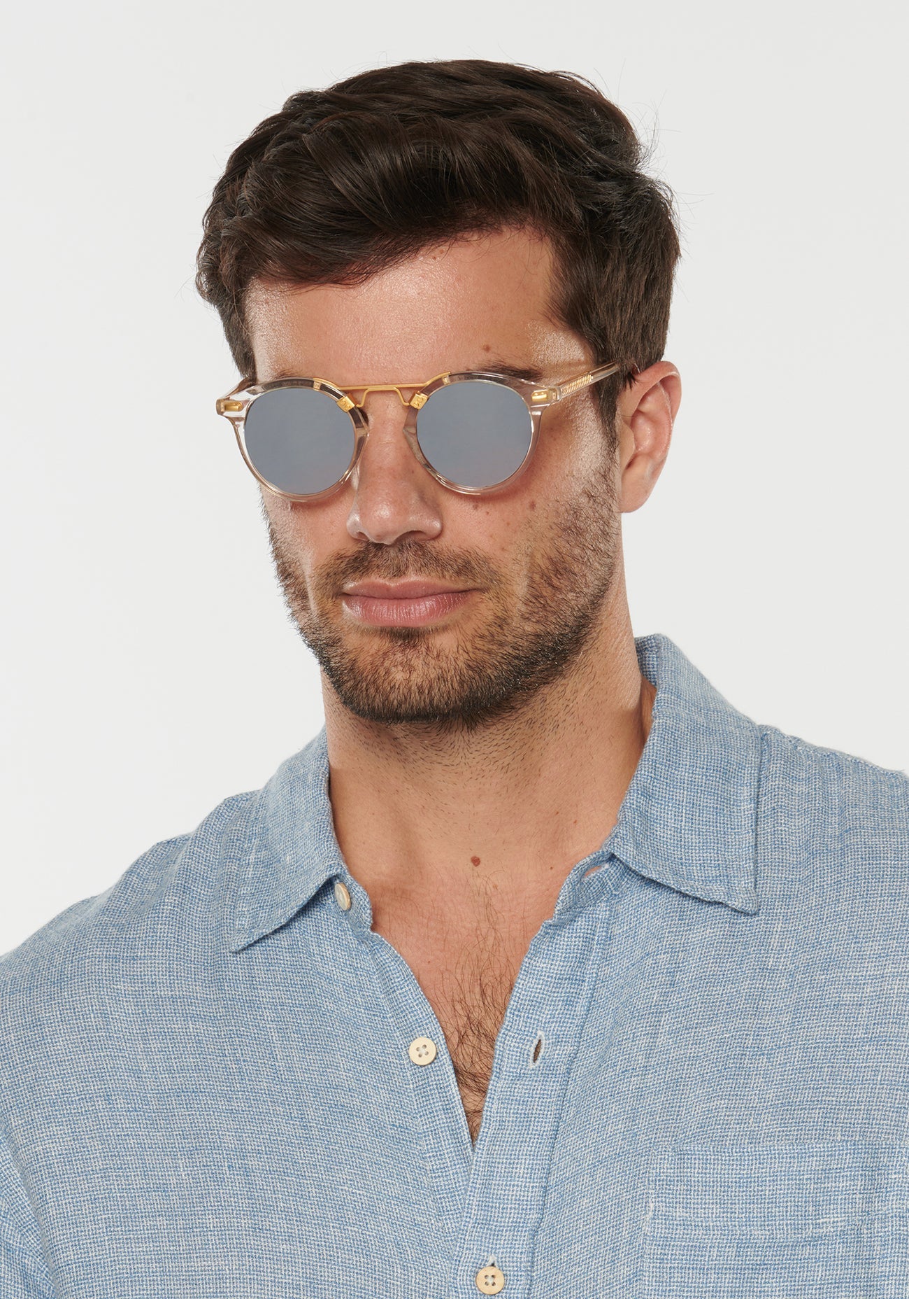 A man wearing Krewe St. Louis Mirrored Sunglasses—iconic classic round gold frames with mirrored lenses—and a light blue button-up shirt looks straight ahead against a plain background.