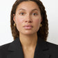 A woman with curly brown hair wears gold-plated Steffie Earrings by Lie Studio and a black blazer, looking straight at the camera against a plain white background.
