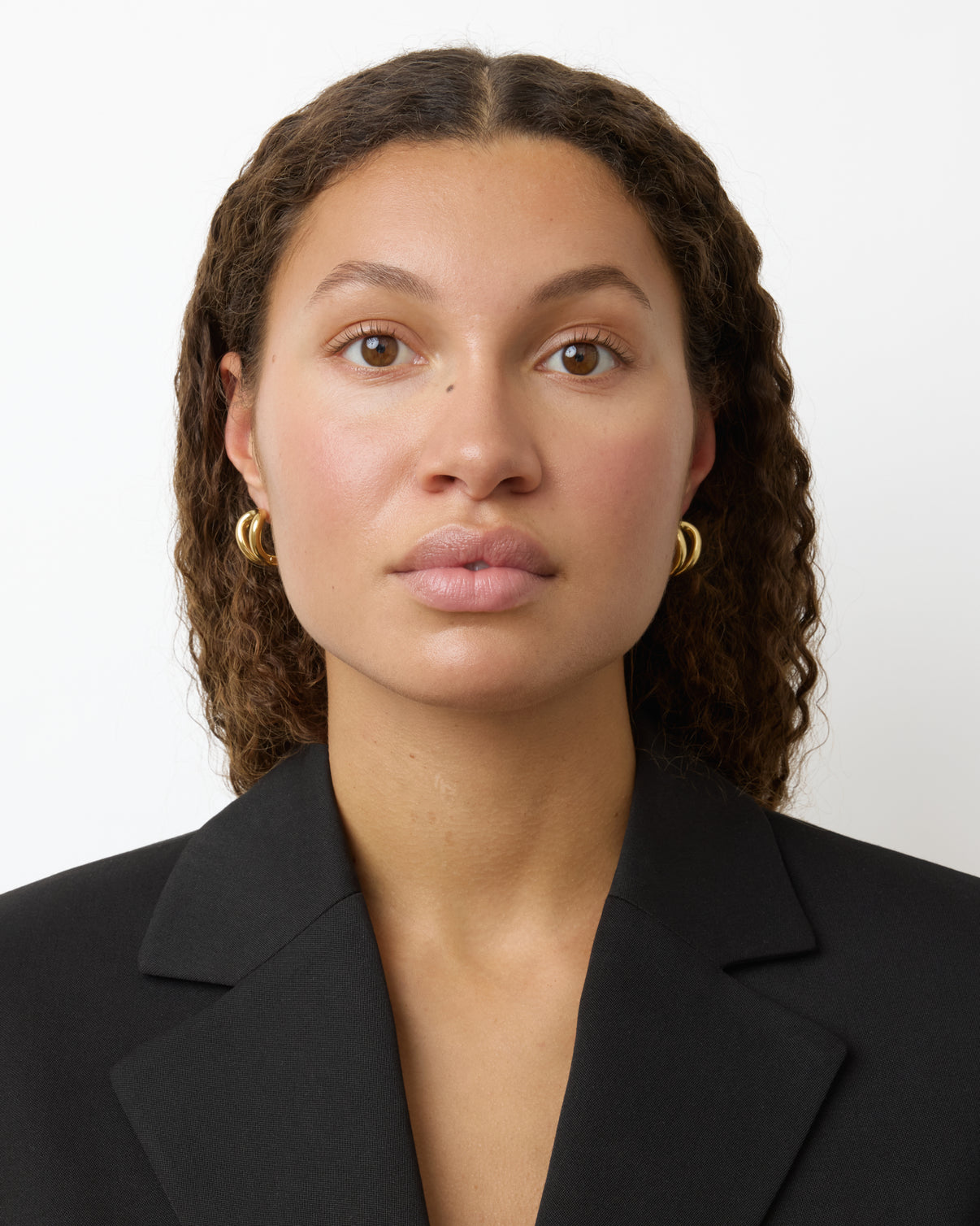 A woman with curly brown hair wears gold-plated Steffie Earrings by Lie Studio and a black blazer, looking straight at the camera against a plain white background.