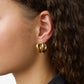 Close-up of a person with curly brown hair wearing a chunky, gold-plated Steffie Earring by Lie Studio in their left ear, paired with a black outfit.