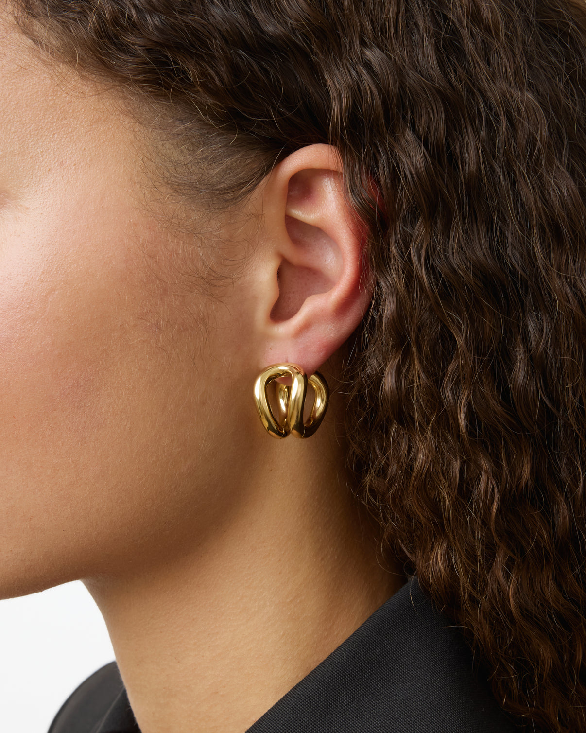 Close-up of a person with curly brown hair wearing a chunky, gold-plated Steffie Earring by Lie Studio in their left ear, paired with a black outfit.