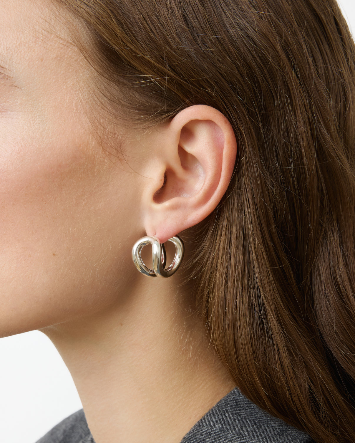 Close-up of a person with brown hair wearing Lie Studio's Steffie Earrings—large, sterling silver double hoops—in their left ear. The individual is dressed in a grey textured garment.