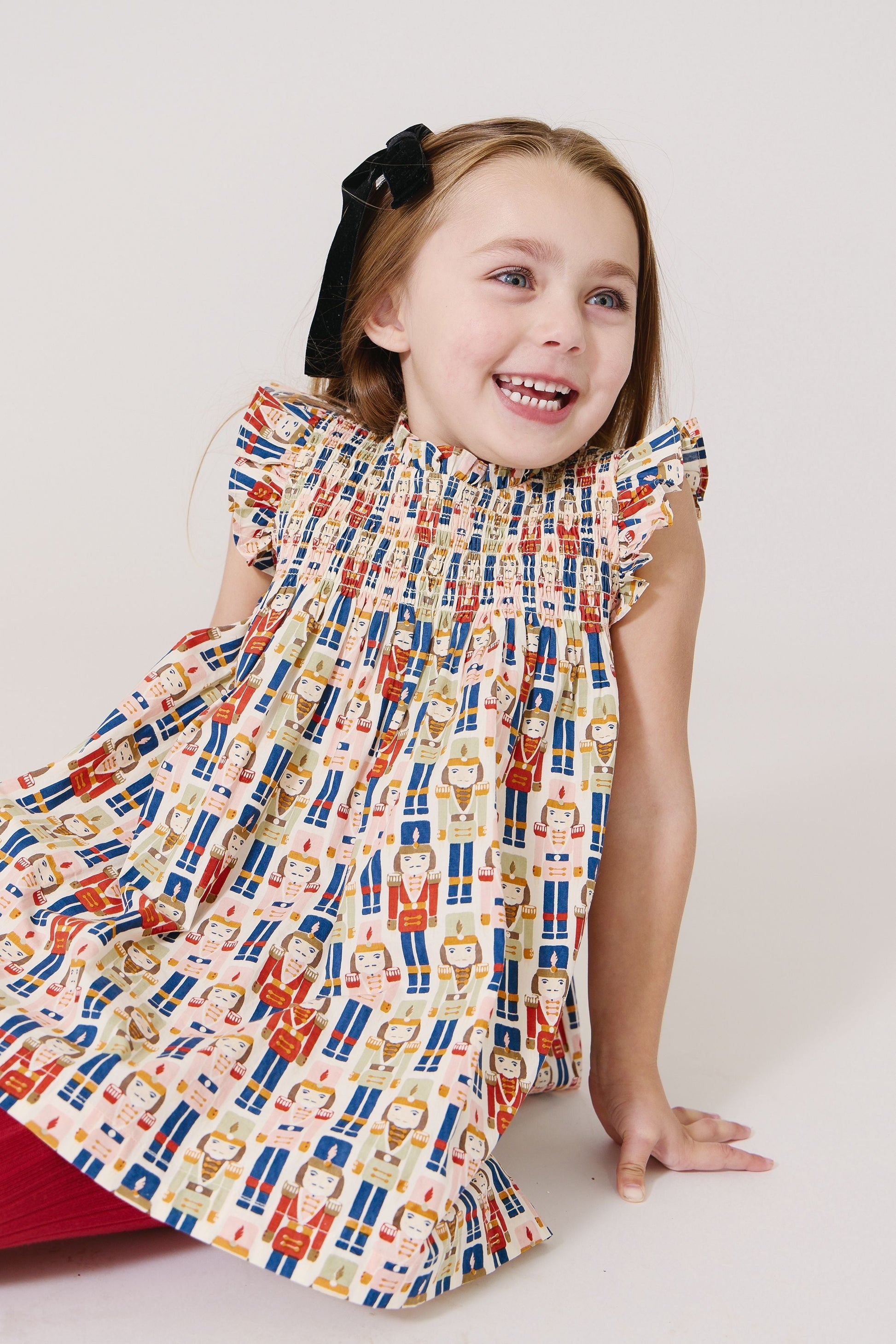 A young girl, wearing a vibrant Pink Chicken Girls Stevie Dress featuring cartoon characters and smocked detailing, is smiling as she sits on the floor with a black hair bow accentuating her look.