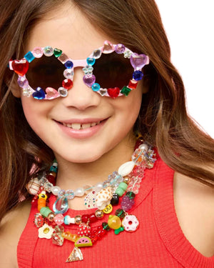 A child beams brightly in a red top, adorned with vibrant bead necklaces and wearing the Super Smalls Sparkle and Shine Gemify Sunglasses and Case. These stylish shades from Super Smalls add flair with their colorful gem stickers while providing essential UVA/UVB protection.