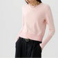 A woman, her face partially out of frame, wears the Sunwash Shrunken Crew by Guest in Residence—a light pink, lightweight cotton-cashmere crewneck sweater with ribbed cuffs—paired with a beaded necklace and black belted trousers. This sweater makes an ideal summer layer.