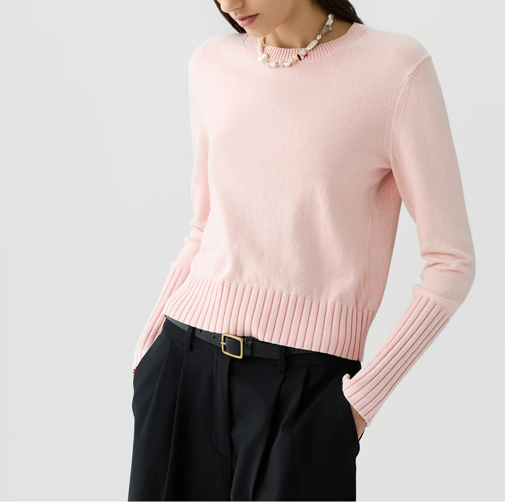 A woman, her face partially out of frame, wears the Sunwash Shrunken Crew by Guest in Residence—a light pink, lightweight cotton-cashmere crewneck sweater with ribbed cuffs—paired with a beaded necklace and black belted trousers. This sweater makes an ideal summer layer.