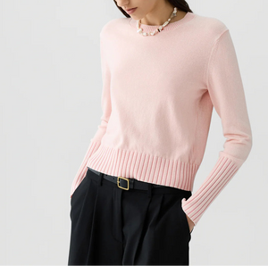 A woman, her face partially out of frame, wears the Sunwash Shrunken Crew by Guest in Residence—a light pink, lightweight cotton-cashmere crewneck sweater with ribbed cuffs—paired with a beaded necklace and black belted trousers. This sweater makes an ideal summer layer.