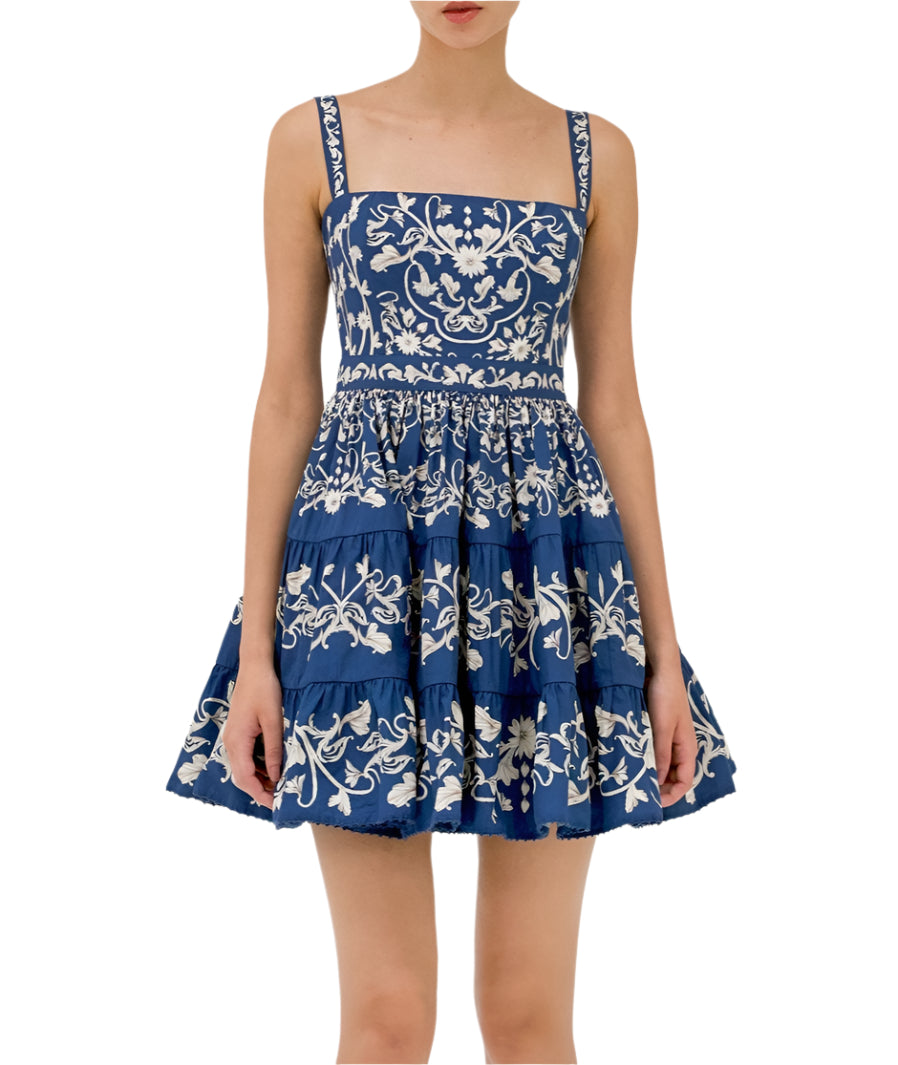 A woman wears the Lima Mini Dress by Agua By Aguabendita, a blue sleeveless cotton mini dress with white floral embroidery and a tiered skirt, standing against a plain background.