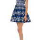 A person wearing the Lima Mini Dress by Agua By Aguabendita—a blue and white, sleeveless, knee-length A-line dress with floral patterns—paired with white sandals, standing against a white background.