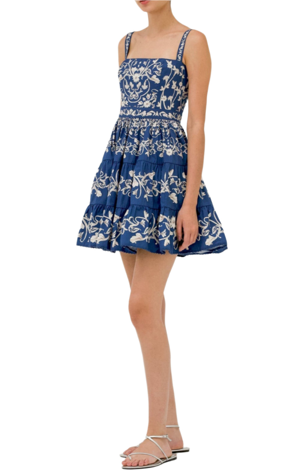 A person wearing the Lima Mini Dress by Agua By Aguabendita—a blue and white, sleeveless, knee-length A-line dress with floral patterns—paired with white sandals, standing against a white background.
