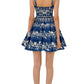 A woman stands with her back to the camera, wearing the Lima Mini Dress by Agua By Aguabendita—a blue sleeveless cotton mini dress featuring white floral embroidery—paired with silver sandals.