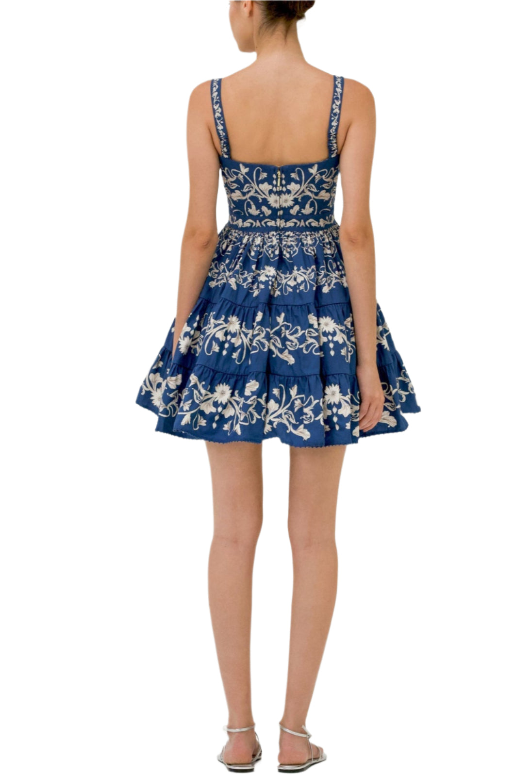 A woman stands with her back to the camera, wearing the Lima Mini Dress by Agua By Aguabendita—a blue sleeveless cotton mini dress featuring white floral embroidery—paired with silver sandals.