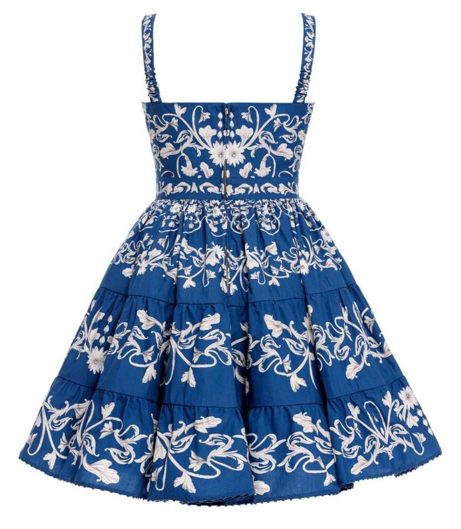 The Lima Mini Dress by Agua By Aguabendita is a blue sleeveless cotton mini dress with a fitted bodice, white floral embroidery, button details, and a tiered skirt, shown from the back.