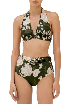 A woman wears the Marimar Top and Vaiven Bottom Bikini by Agua By Aguabendita, a dark green recycled polyester set with large white flowers, halter top, high-waisted bottoms, and UPF 50 sun protection.