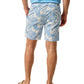 A person wearing Johnnie-O Salty Dogs Swim Trunks in blue with fun prints, a light blue shirt, and beige flip-flops stands with hands in pockets, facing away.