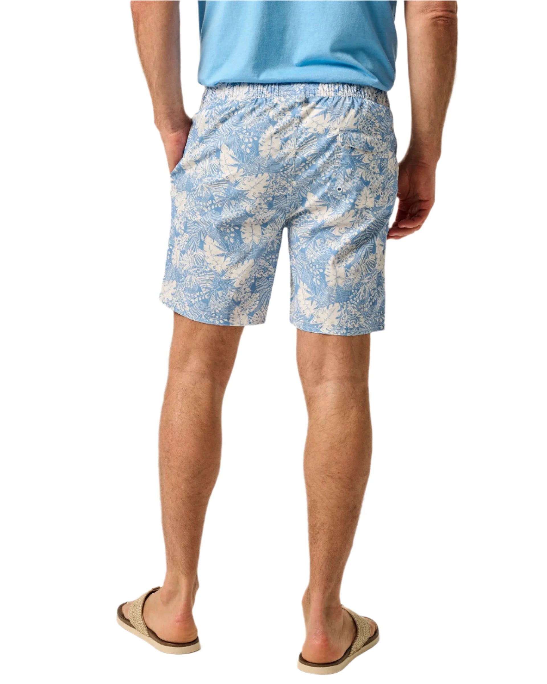 A person wearing Johnnie-O Salty Dogs Swim Trunks in blue with fun prints, a light blue shirt, and beige flip-flops stands with hands in pockets, facing away.