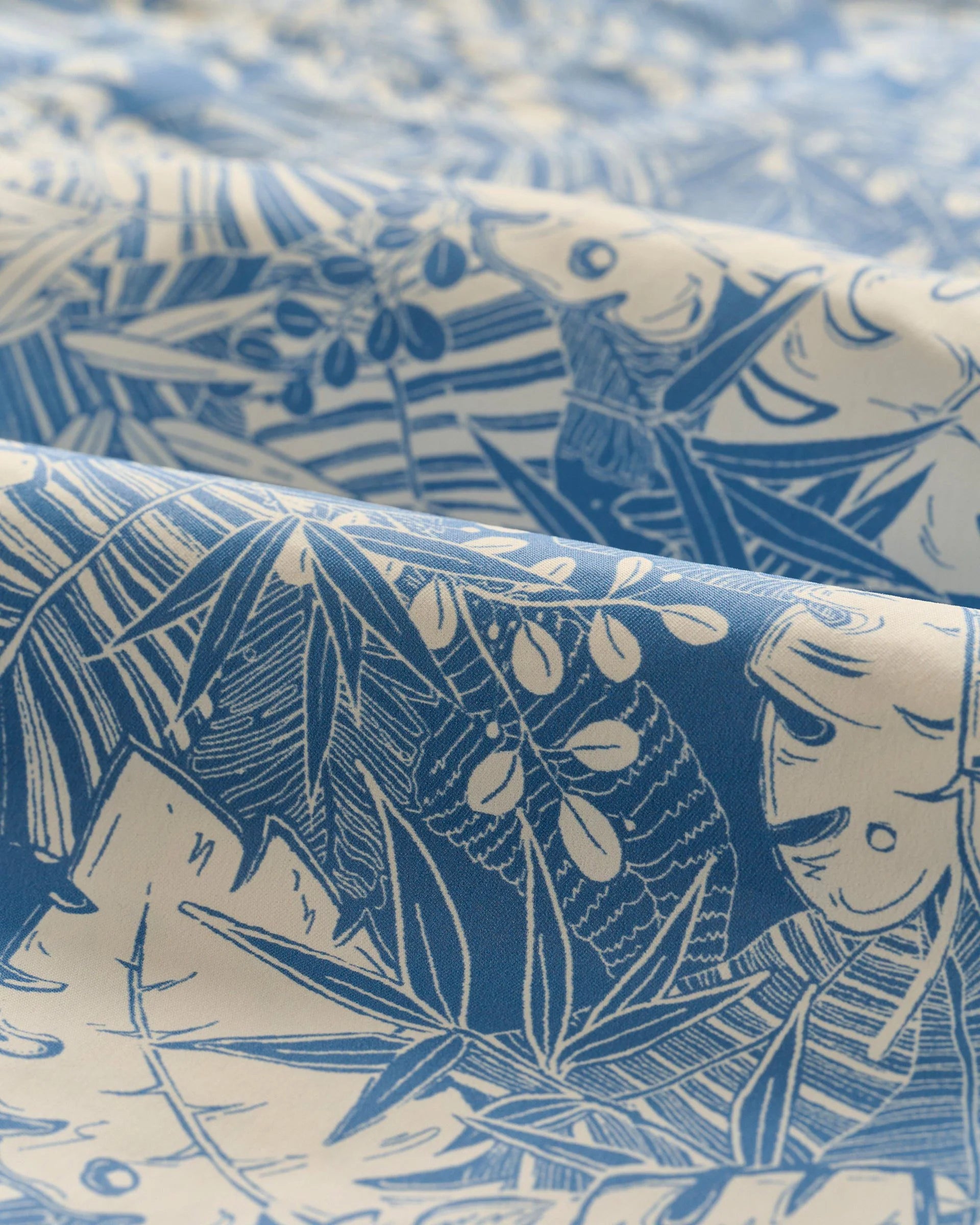 Close-up of the Johnnie-O Salty Dogs Swim Trunks showcases a blue and white fabric with fun tropical leaf and plant patterns, gently draped and folded—ideal for crafting comfortable surf shorts by Johnnie-O.