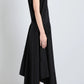 A woman stands in profile wearing the Proenza Schouler Sybella Dress, a sleeveless black asymmetrical design with a handkerchief hem and fluid silhouette, paired with black strappy sandals against a plain light background.