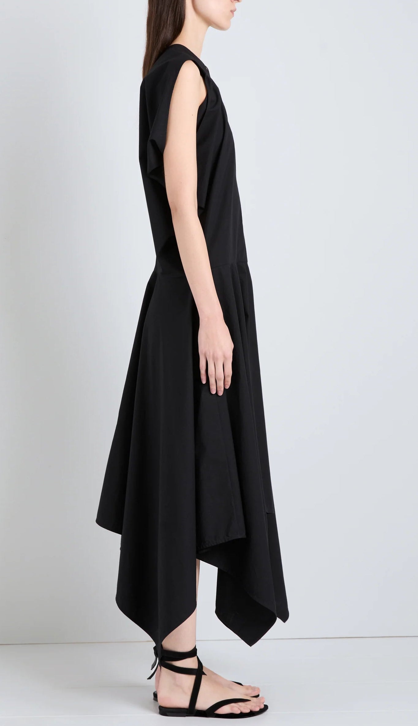 A woman stands in profile wearing the Proenza Schouler Sybella Dress, a sleeveless black asymmetrical design with a handkerchief hem and fluid silhouette, paired with black strappy sandals against a plain light background.