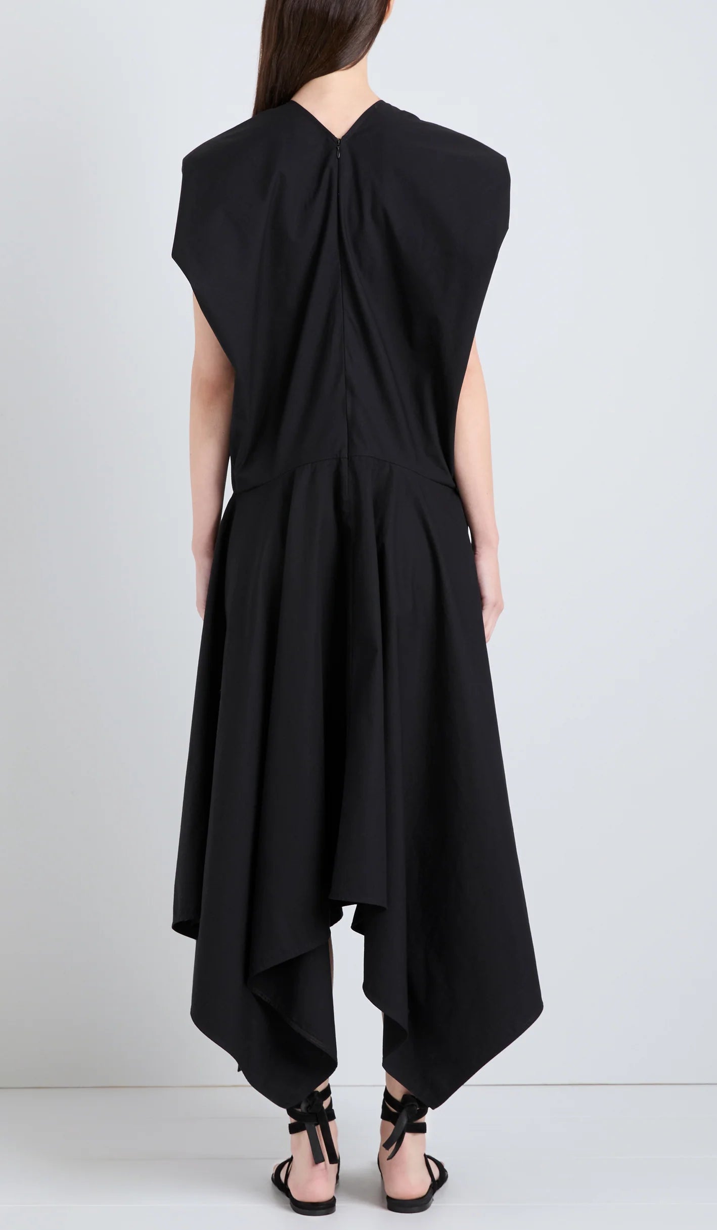 A person is seen from the back wearing the Sybella Dress by Proenza Schouler, a sleeveless black design featuring a handkerchief hem and flowing silhouette, set against a plain light background.