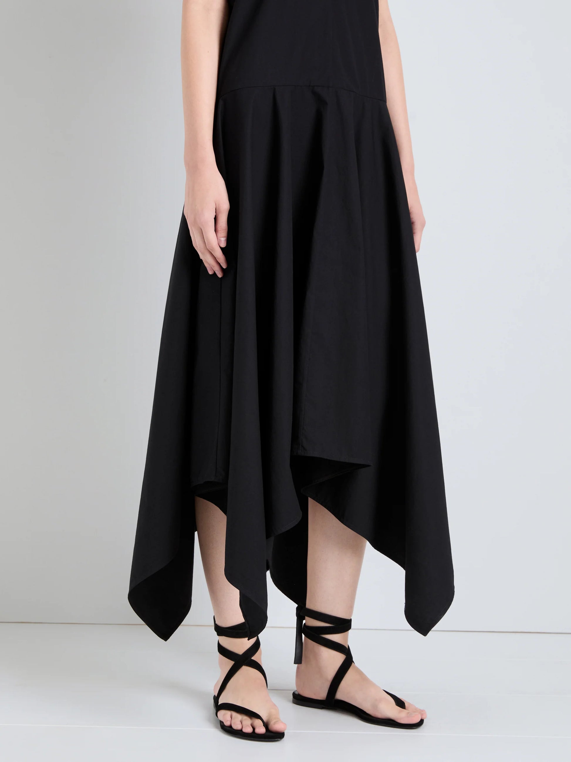 A person wearing the Sybella Dress by Proenza Schouler, featuring a black handkerchief hem and flowing silhouette, pairs it with black strappy sandals and stands against a plain light-colored background.