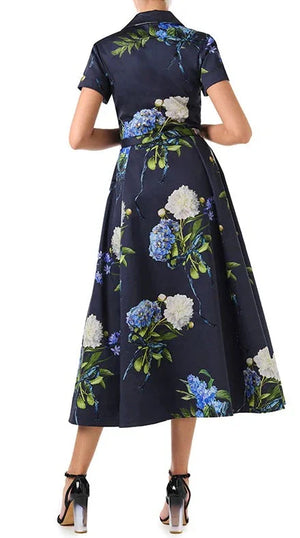 A person wearing a Monique Lhuillier Short Sleeve A Line Printed Poplin Dress with a collar and belted waist. The dress, adorned with blue and white flowers, falls to midi length. They are also wearing black heels.