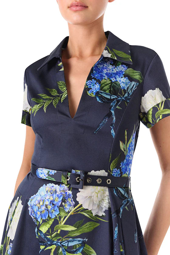 A person wearing a Monique Lhuillier Short Sleeve A Line Printed Poplin Dress by Monique Lhuillier with a deep V-neck, short sleeves, and a floral print featuring blue and white flowers. The belted waist adds a stylish touch to the ensemble.