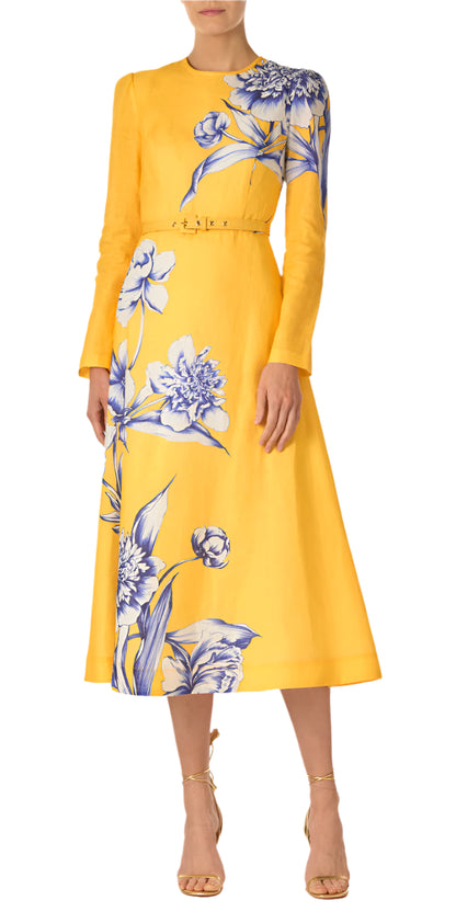 A person wearing the Monique Lhuillier Long Sleeve Floral Day Dress, featuring a jewel neckline, vibrant yellow with blue floral patterns, and a gold belt, paired with tan strappy heels.